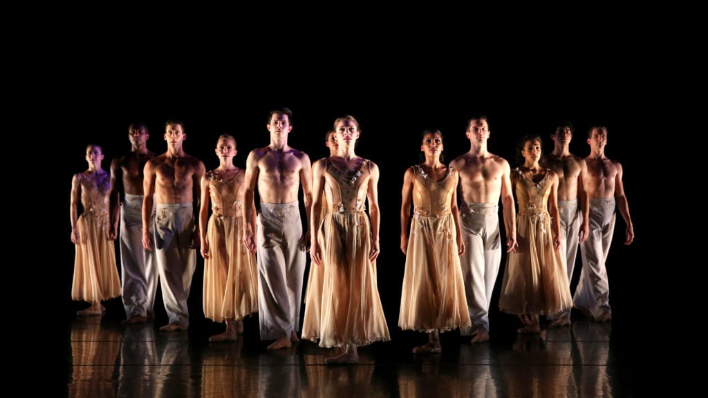 A group of dancers stand with arms by their side in dramatic lighting.